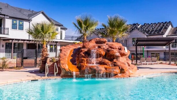 Une piscine agrémentée d’une cascade artificielle en roches, entourée de palmiers et de maisons modernes.