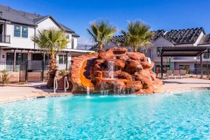 A swimming pool with a rock waterfall feature, surrounded by palm trees and modern houses.
