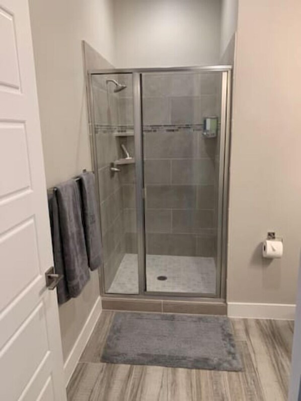 A walk-in shower with a glass door, a towel rack with towels, a toilet paper holder, and a gray rug on a wooden floor.