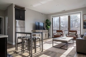 A modern living room with a large window, a kitchen with dark cabinets, a flat-screen TV, and a dining area with stools.