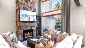 A living room with a stone fireplace, a television mounted above it, and a view of the outdoors through large windows.