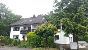 A white building with a balcony, surrounded by greenery and a paved area with a bench. - Ferienwohnung am Ententeich (Bad Marienberg)