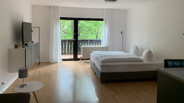 A modern bedroom with a bed, a TV, a desk, and a balcony with a view of greenery. - Ferienwohnung am Ententeich (Bad Marienberg)