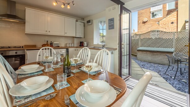 Un coin repas avec une table en bois dressée pour six personnes, des chaises blanches et une vue sur le jardin à travers une porte vitrée.