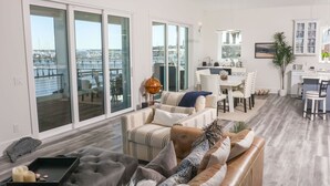 Uma sala de estar ampla com vista para um porto, uma área de jantar com mesa e cadeiras brancas, e uma cozinha com armários brancos.