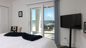 Quarto de hotel com cama, televisão, quadro na parede e vista para o mar por uma porta de vidro deslizante.