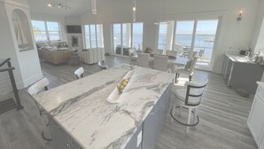 Uma cozinha espaçosa com bancada de mármore, grandes janelas com vista para a água e uma área de refeições com mobiliário branco.