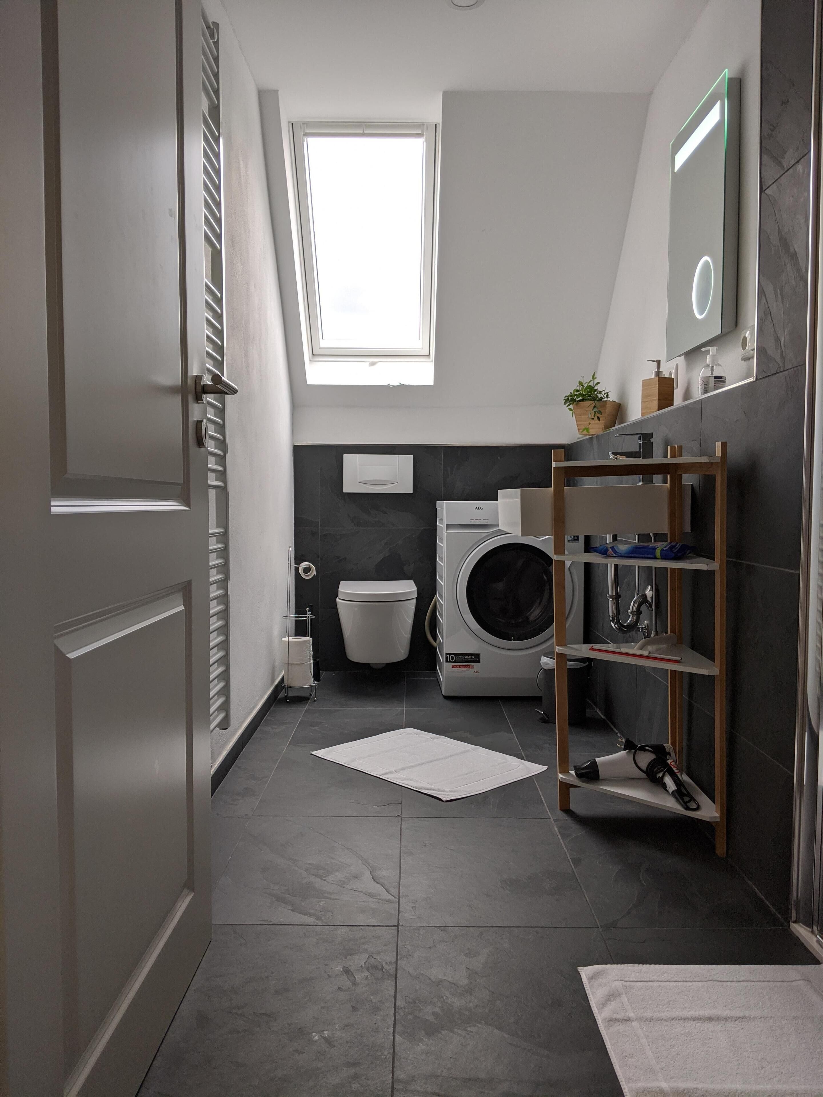 Un baño moderno con tragaluz, lavadora, inodoro y estante de madera con artículos de aseo.