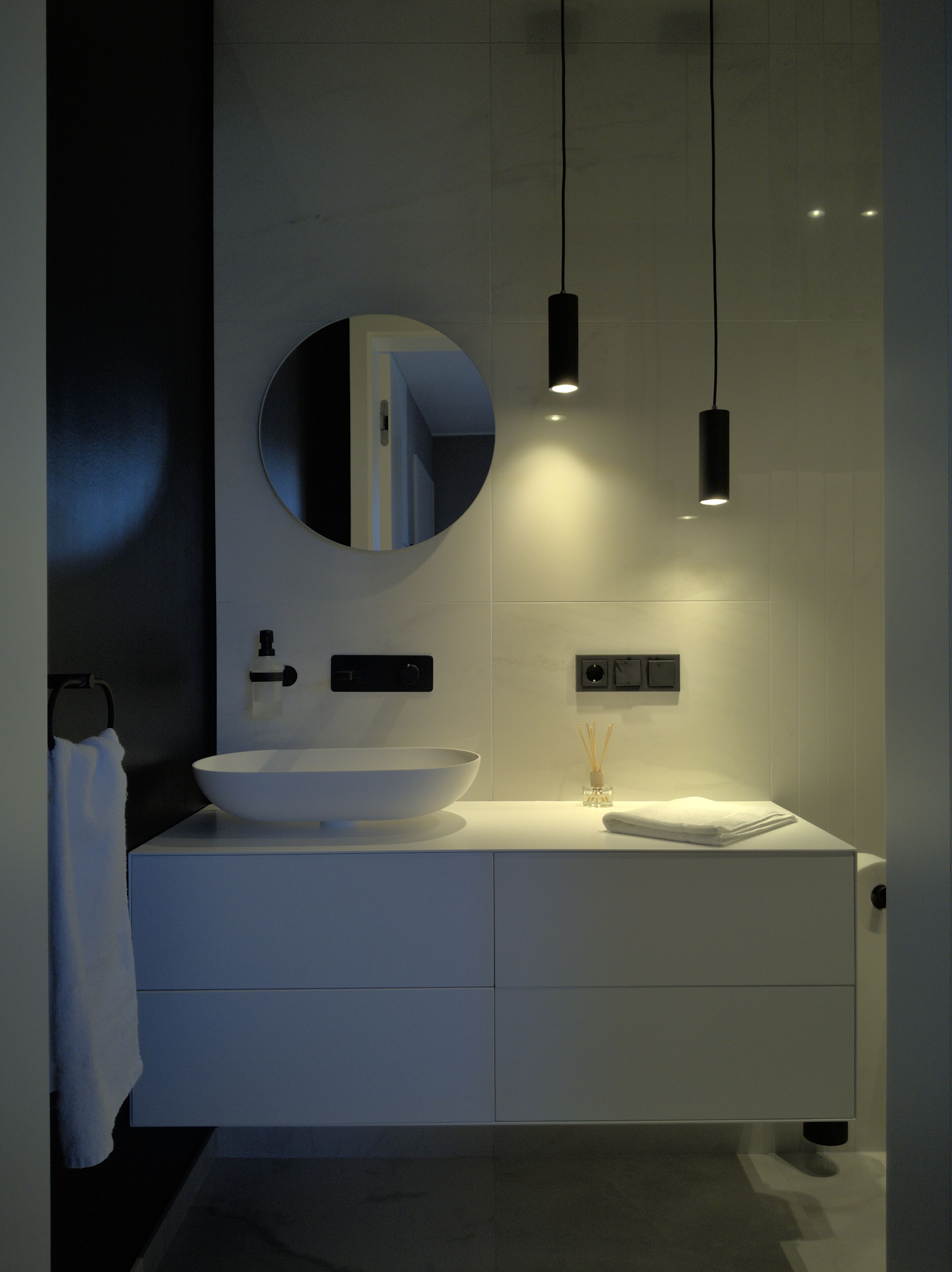 A modern bathroom with a white sink, a round mirror, and pendant lights.