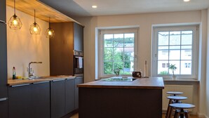 Een moderne keuken met donkere kasten, een centraal eiland, hanglampen en uitzicht op de omgeving door ramen.