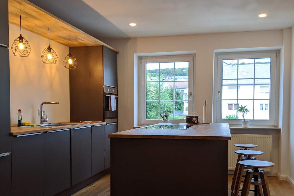 Une cuisine moderne avec des meubles de couleur sombre, un îlot central, des luminaires suspendus et une vue sur l’extérieur grâce à de grandes fenêtres.