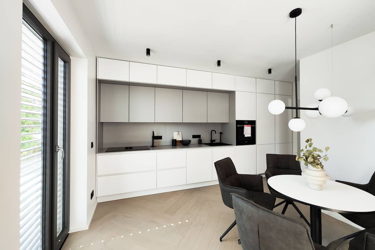 Une cuisine moderne avec des meubles blancs, un plan de travail noir et un four encastré. On y trouve un coin repas avec une table ronde et des chaises, ainsi qu’un luminaire suspendu au-dessus de la table.