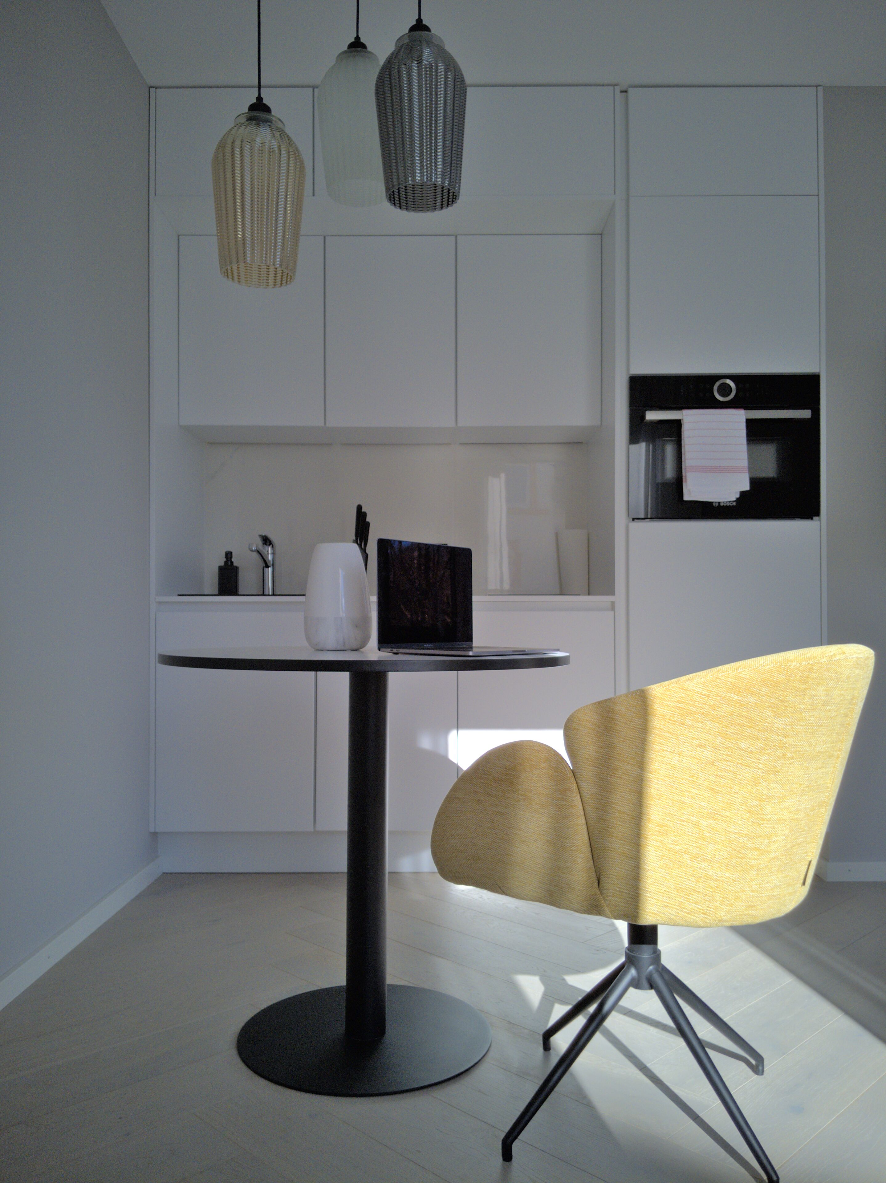 A modern kitchen with white cabinets, a round table, a yellow chair, and pendant lights.