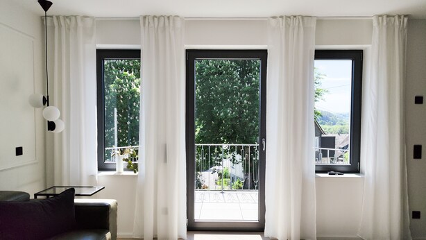 Een moderne woonkamer met een zwarte leren bank, een klein tafeltje en een groot raam met witte gordijnen.