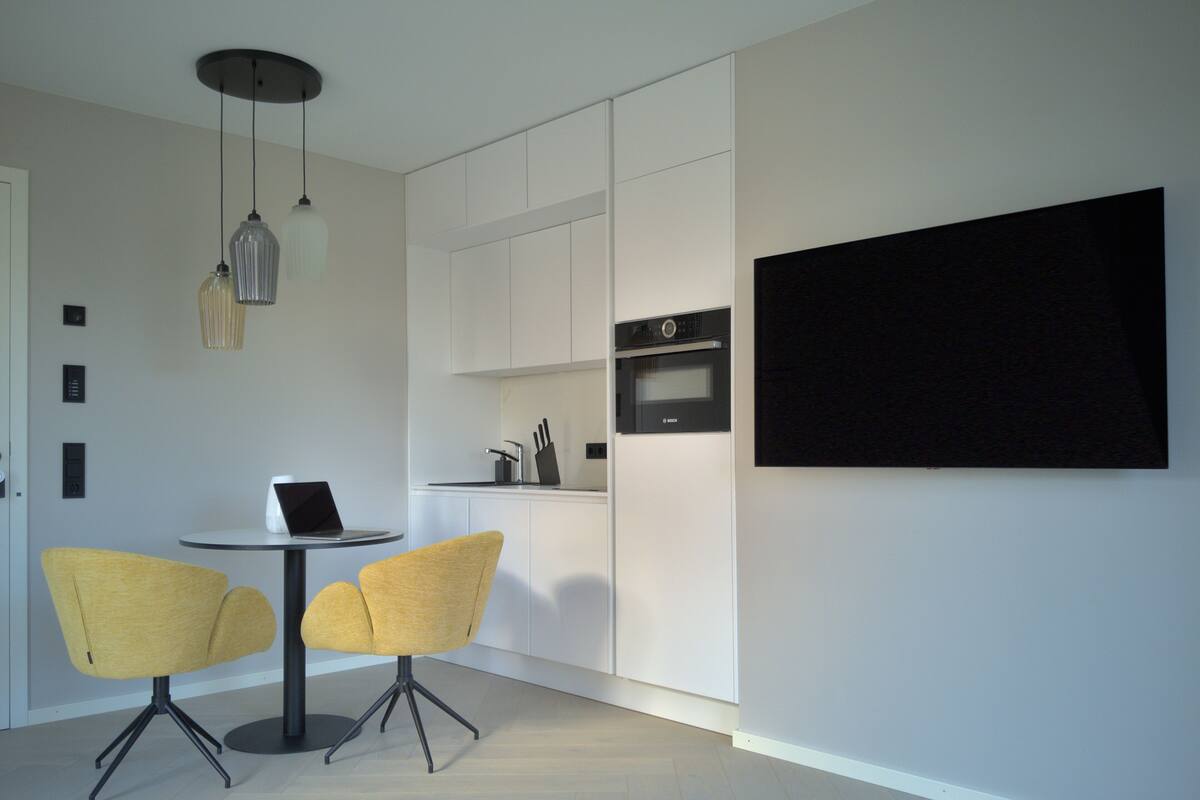 Une cuisine moderne avec des meubles blancs, un four Bosch et un coin repas comprenant deux chaises jaunes et un ordinateur portable posé sur une petite table.