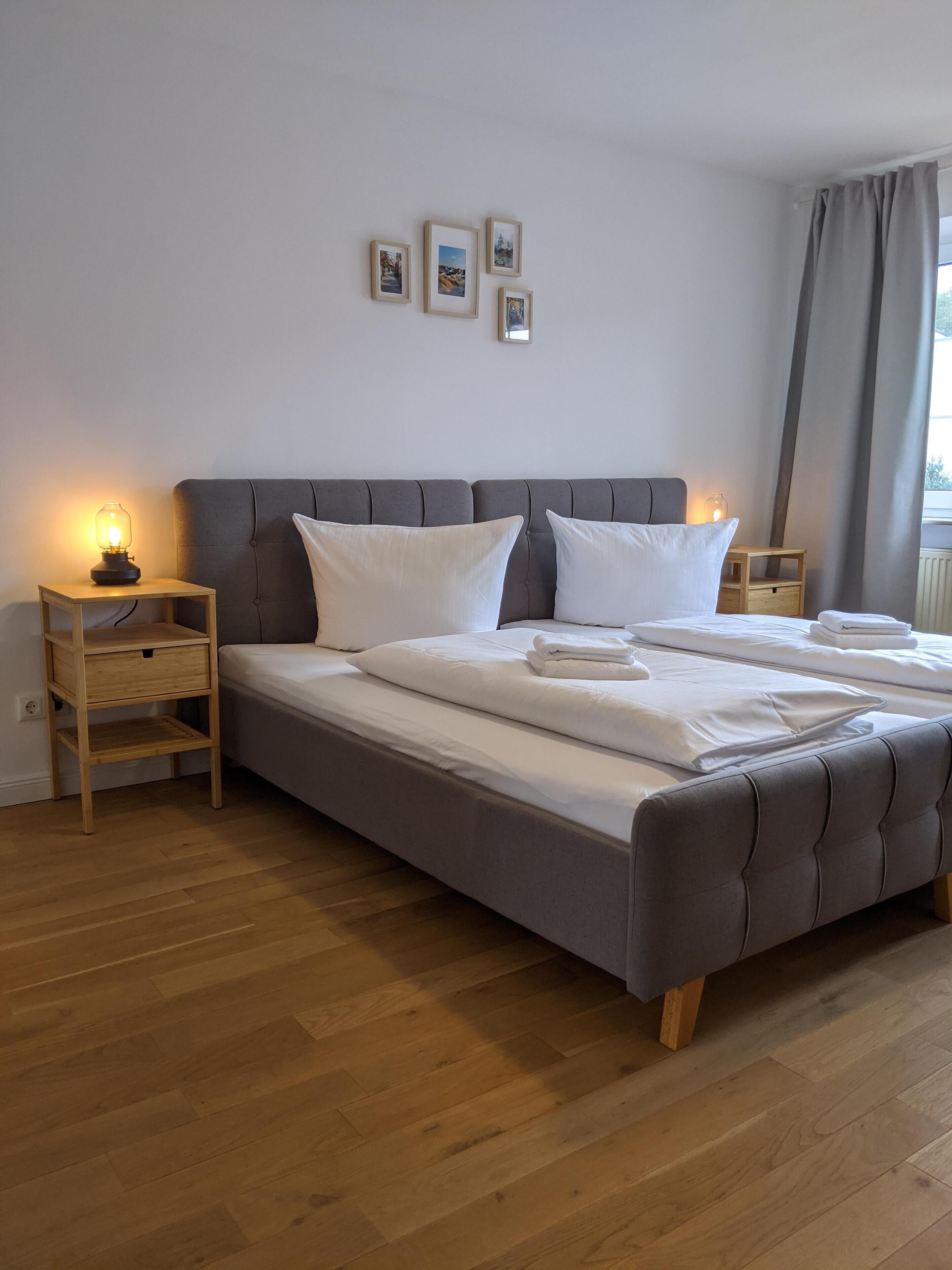 Una cama bien hecha con sábanas blancas, dos mesitas de noche con lámparas, un cabecero tapizado en gris y un suelo de madera.