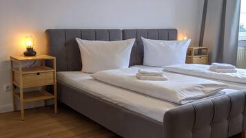 Una cama bien hecha con sábanas blancas, dos mesitas de noche con lámparas, un cabecero tapizado en gris y un suelo de madera.