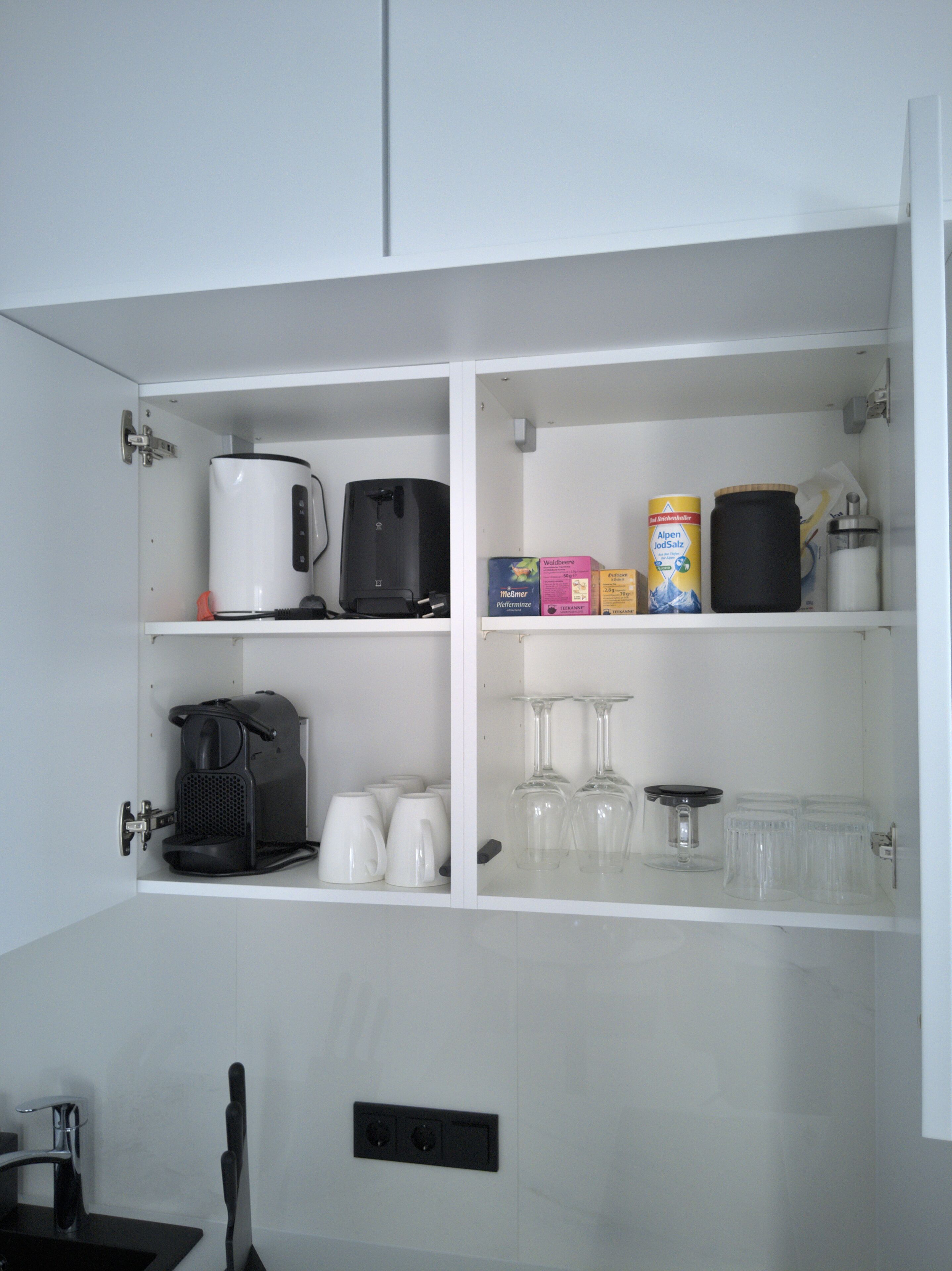 A kitchen with a coffee maker, a kettle, a box of coffee pods, a container of coffee creamer, and a set of wine glasses.