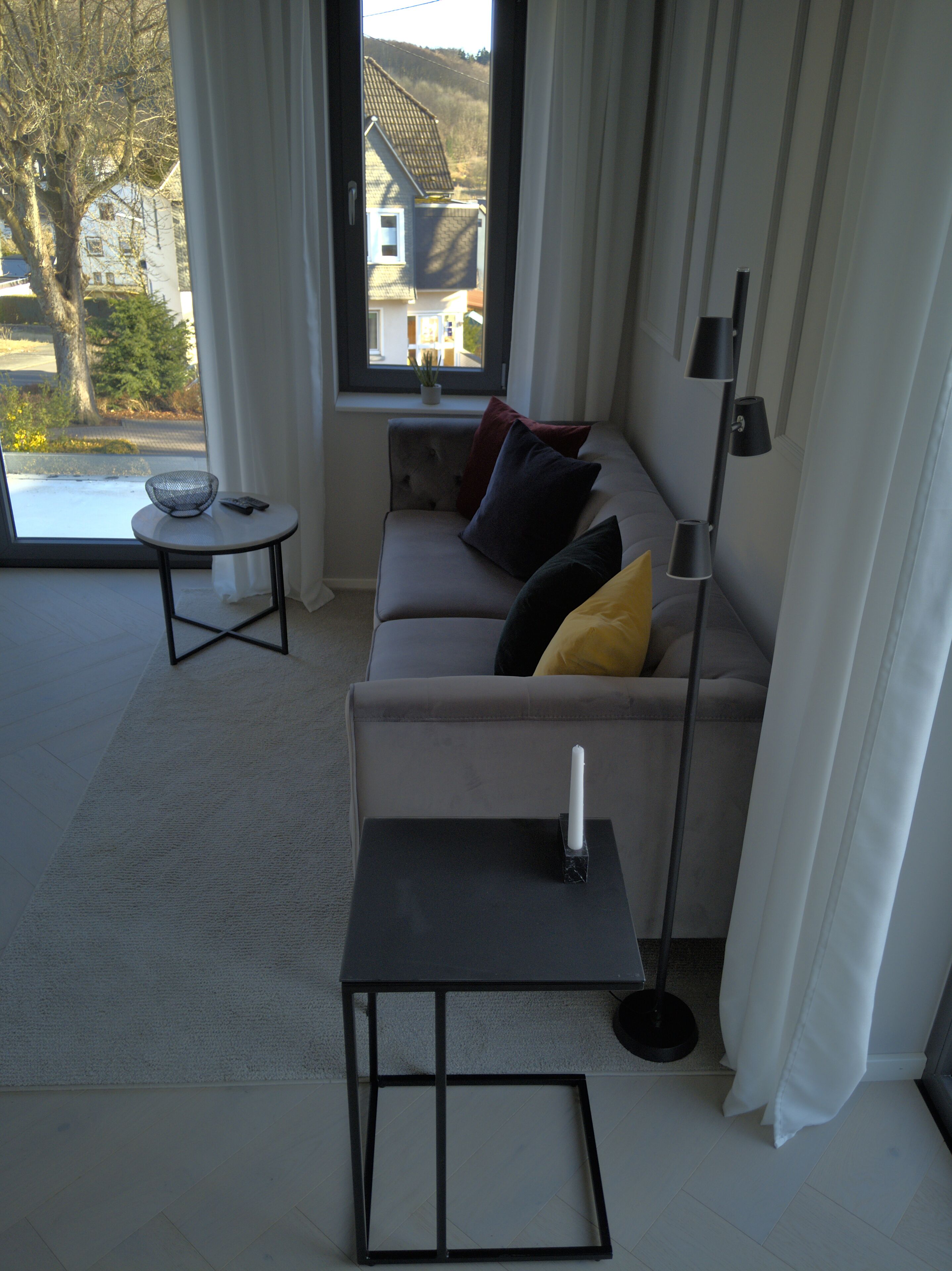 A modern living room with a sofa, a small table, and a floor lamp. A window provides a view of the outside.