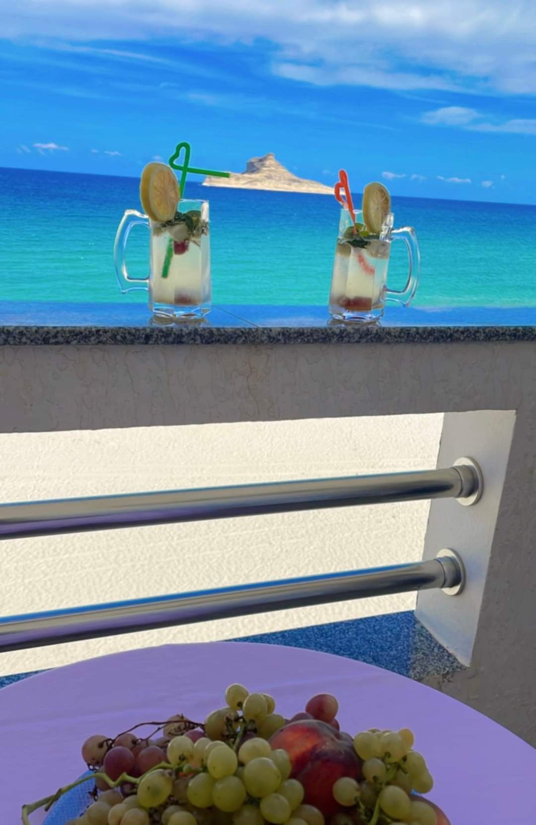 Deux boissons sur un balcon avec vue sur l’océan et une table avec des raisins.