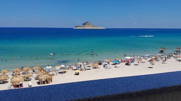 Une plage avec des parasols en chaume et des chaises longues, une mer d’un bleu limpide et une île qui se dessine à l’horizon.