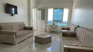A spacious living room with a large window offering a view of the sea, a flat-screen TV mounted on the wall, and a dining area with a table and chairs. - S+1 h standing (Rafraf)