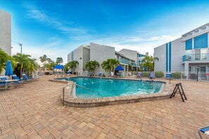 A swimming pool area with lounge chairs and umbrellas, surrounded by palm trees and a building with balconies. - Pickleball, Pools & Docks - Yacht Club #165A (Madeira Beach)