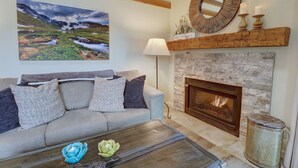 A living room with a fireplace, a sofa with cushions, a coffee table with decorative items, and a painting of a landscape on the wall. - NEW! Mountain Condo, Outdoor Hot Tubs, Stunning Summer Views - 201C (Frisco)