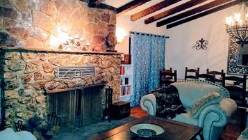 A cozy living room with a stone fireplace, wooden beams, a white sofa, and a dining area with a chandelier.