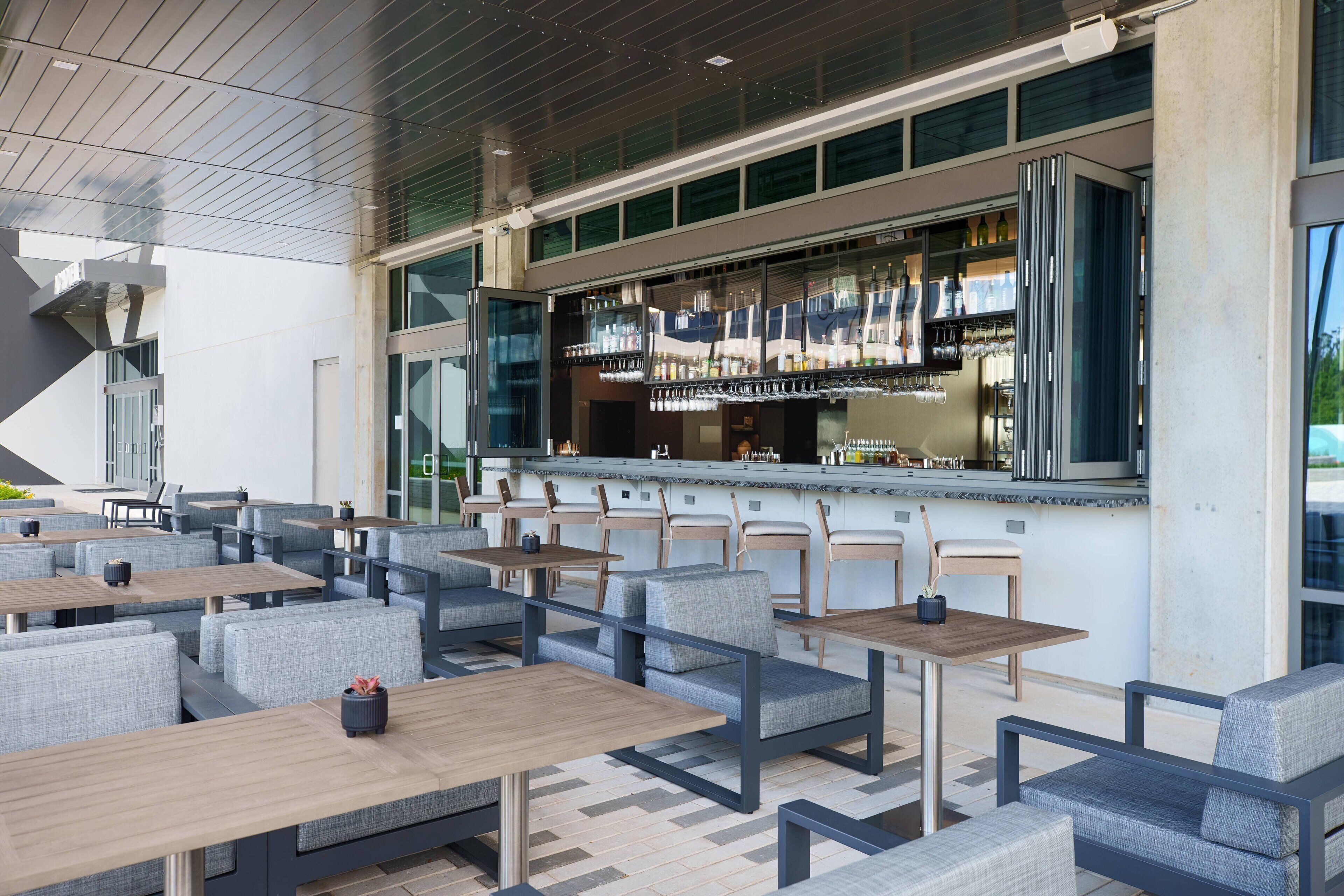 Un espace extérieur moderne aménagé avec des tables et des chaises, un bar doté d’une paroi vitrée, et offrant une vue sur l’extérieur.
