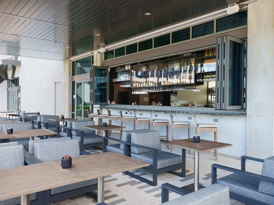 Un espace extérieur moderne aménagé avec des tables et des chaises, un bar doté d’une paroi vitrée, et offrant une vue sur l’extérieur.