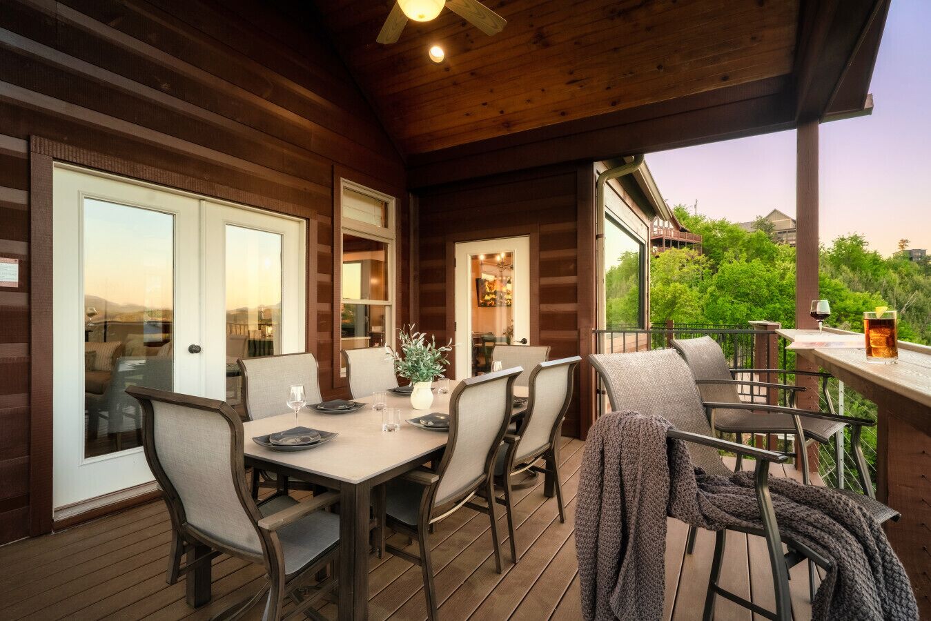 A wooden deck with a table and chairs, a glass of drink, and a view of greenery.