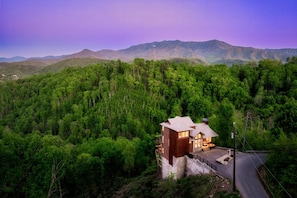 A two-story wooden cabin with a stone foundation, surrounded by dense forest and mountains in the background.