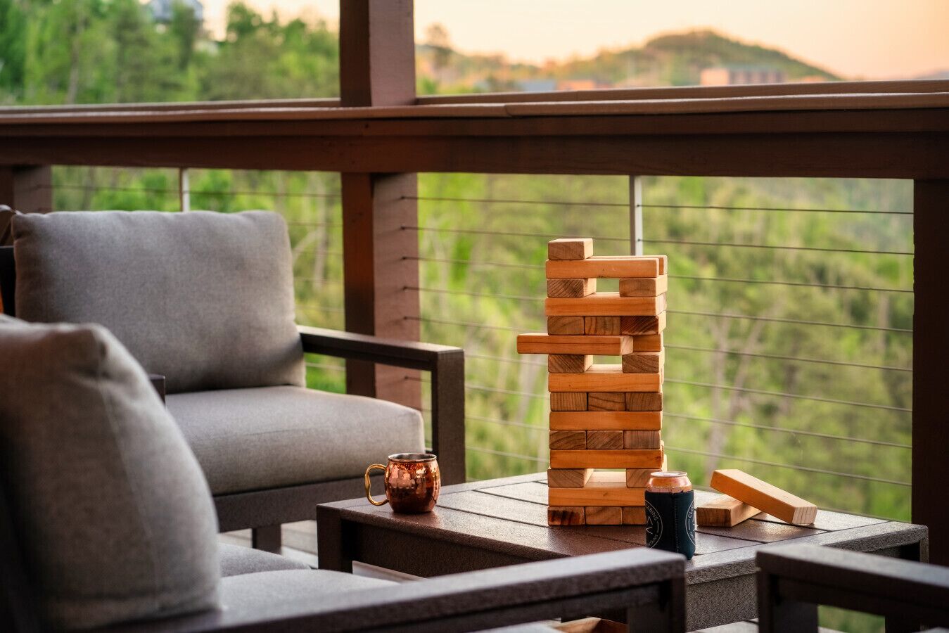 Una torre de Jenga de madera sobre una mesa, un vaso de cobre y una silla con vista a la vegetación exterior.