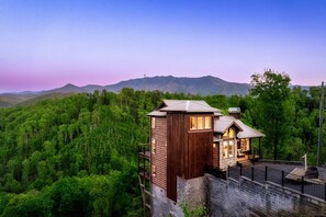 A wooden cabin with a balcony, surrounded by dense forest and mountains.