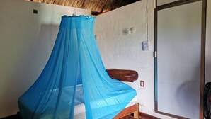 Una habitación con una cama bajo un dosel azul, suelo de madera, una puerta y una lámpara fijada a la pared.