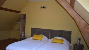 A bedroom with a wooden ceiling, a bed with white bedding and two yellow pillows, a nightstand with a lamp, and a shelf with decorative items.