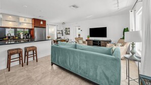 A modern living room with a teal sofa, wooden bar stools, a kitchen with a stainless steel refrigerator, and a flat-screen TV mounted on the wall. - 1 BR - Guesthouse - A beachside gem! Walk 1 min to Beach! (Boca Raton)
