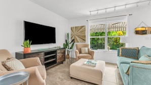 A modern living room with a flat-screen TV, comfortable seating, a coffee table, and a large window with a view of greenery. - 1 BR - Guesthouse - A beachside gem! Walk 1 min to Beach! (Boca Raton)