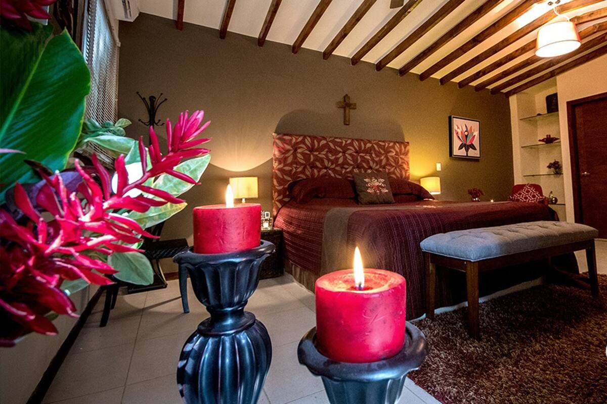 A bedroom with a bed, two red candles on black stands, a cross on the wall, and a bench.