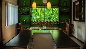 A dining area with a long table, chairs, hanging lights, and a window with plants.