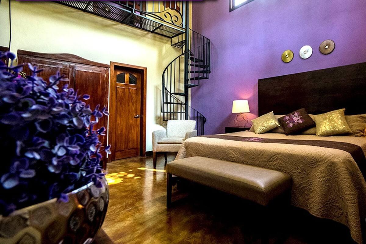 A bedroom with a bed, a chair, a small table, a lamp, and a staircase.