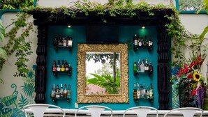 An outdoor seating area with turquoise chairs and a small tree centerpiece, surrounded by a teal and white wall with a cross and various bottles displayed.