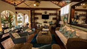 A spacious living room with a large sofa, a coffee table, and a television. There are multiple seating options, including a chair and a couch. The room has a wooden ceiling and a large window.