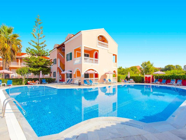 A swimming pool with lounge chairs and umbrellas, surrounded by a building with balconies and a clear blue sky.
