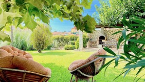 Two wicker chairs with cushions in a grassy garden area with a stone wall and a stone oven in the background. - Asteraki Beach House (Acharavi)