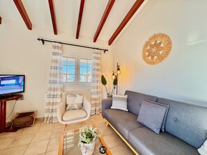 Ein Wohnzimmer mit einer Couch, einem Sessel, einem Fernseher, einem Fenster mit Vorhängen, einer Wanddekoration und einer Stehlampe.