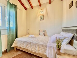A bedroom with a bed, a window with curtains, a wall-mounted light, and a small picture. - Asteraki Poolside Apartment (St. Stefanos Bay)