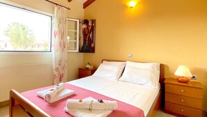 Une chambre d’hôtel avec un lit, une table de chevet avec une lampe, une fenêtre avec des rideaux, un tableau au mur et une vue sur les palmiers à l’extérieur.