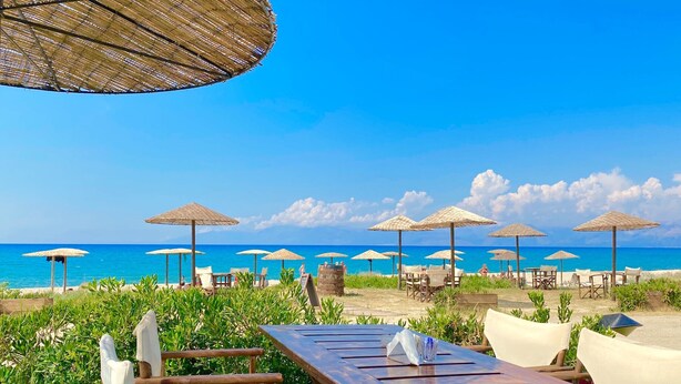 Un restaurant situé en bord de mer, avec des tables et des chaises en bois, entouré de parasols en chaume, et offrant une vue sur l’océan.
